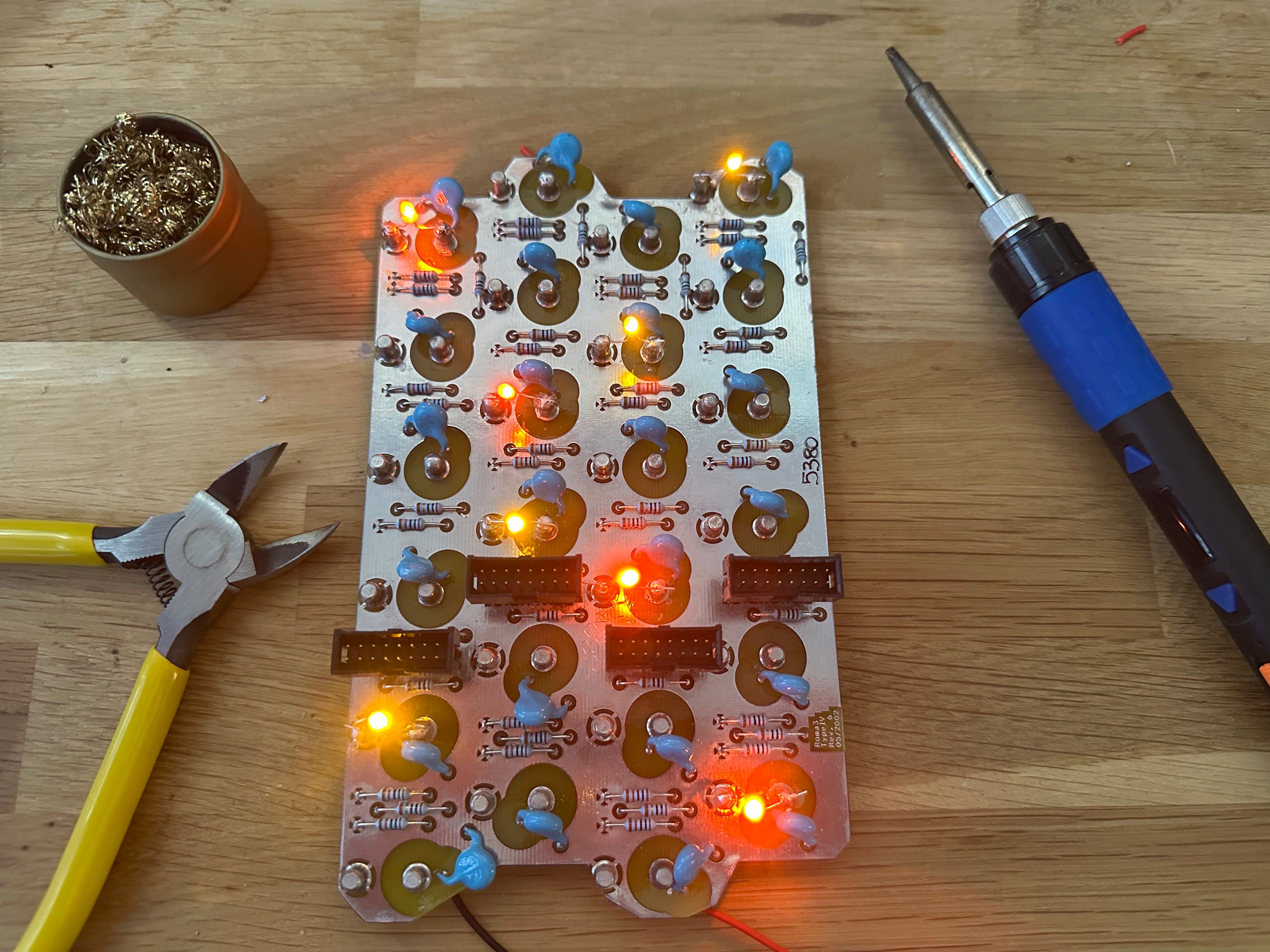 A partially assembled electronic circuit board on a wooden workbench, with several red and yellow LEDs lit up. The board is populated with components such as resistors, capacitors, and headers. To the left is a pair of yellow-handled wire cutters and a brass wire tip cleaner in a small container. To the right is a soldering iron with a blue and black handle. The scene suggests an ongoing electronics assembly or testing project.