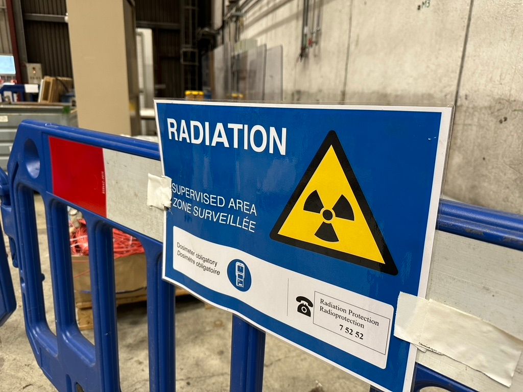 A warning sign attached to a blue safety barrier in an industrial setting, indicating a radiation supervised area. The sign features a prominent yellow and black radiation symbol, with text in English and French stating 'RADIATION – SUPERVISED AREA – ZONE SURVEILLÉE'. It also notes that a dosimeter is obligatory and provides a contact number for Radiation Protection services. The background shows one of the CERN warehouses with equipment and concrete walls.