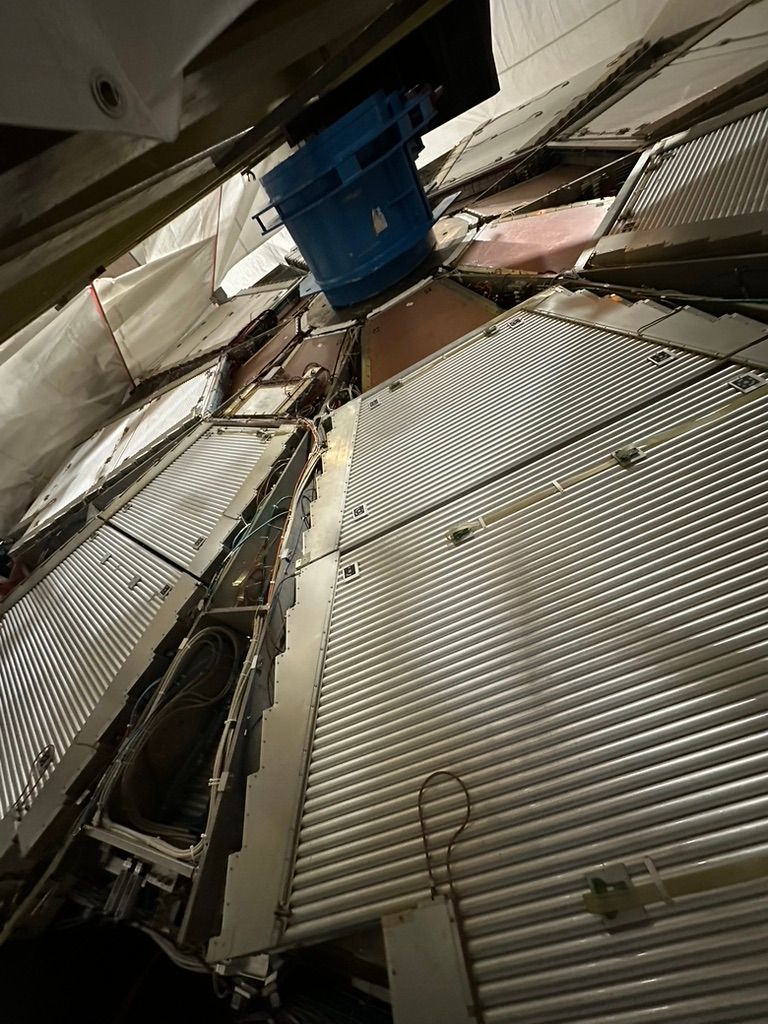 Interior view of a large, segmented detector component. The structure is composed of metallic panels with cooling fins or shielding, arranged radially around a central blue cylindrical hub. Cables and connectors are visible between the segments, suggesting complex instrumentation and data acquisition systems. The scene is enclosed by a protective white tent-like structure