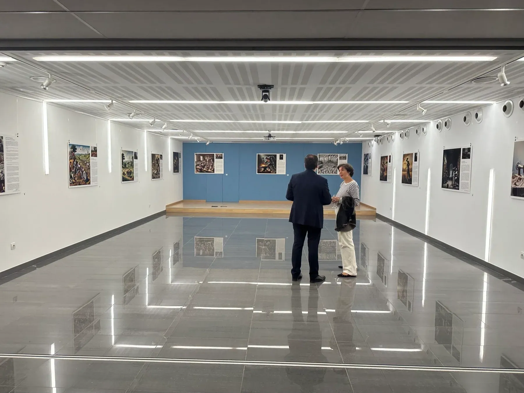 Visitors in a bright gallery with the exhibition posters arranged around the walls.